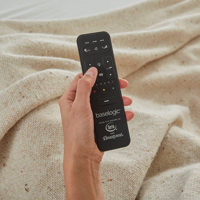 A person holding a Beautyrest wireless remote control while lying on a mattress, with a cozy cream knit blanket and teal bedding, highlighting the ease of adjusting the bed frame's head and foot positions.